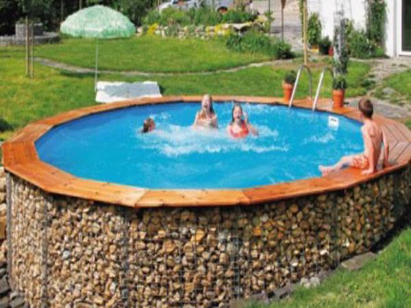 A round gabion swimming pool with 3 people swimming in it and one sitting on it.