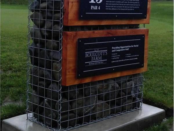 A gabion basket is filled with rocks with two boards on as a sign.