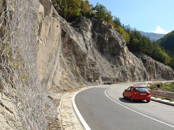 A red car is moving at the foot of a mountain which is protected by hexagonal wire mesh.