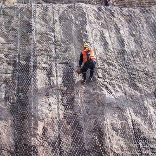 Two workers are installing hexagonal wire mesh rockfall barriers.